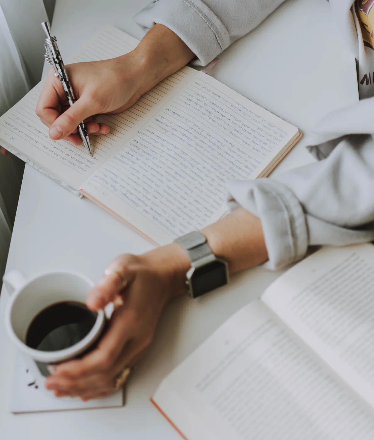Person writing in a notebook beside coffee and open reading material.